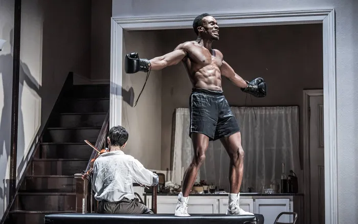 A boxer in gloves and shorts stands framed in a doorway while a violinist plays on the stairs, Golden Boy at the Almeida Theatre